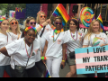 Nurses in white scrubs with rainbow flags and accessories; one carries a sign "I Love My Trans Patients" with the trans flag (pastel blue, pink, and white stripes)