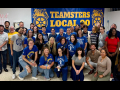 A group of 25 blue-shirted nurses pose happily in front of a Teamsters Local 90 banner
