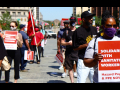 Masked people stand outside holding red printed signs. Visible sign in foreground says: "Solidarity with Sanitation Workers! Hazard Pay and PPE Now!"