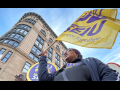 From a heroic low angle, we look up at a Black woman marching in a purple SEIU BJ shirt. The yellow SEIU flag she carries is billowing in the wind, mirroring the curve of the building behind her, against the blue sky. We can see the corners of other picket signs--she is part of a dense crowd.