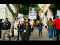 A woman holds a bullhorn and a picket sign that says "On Strike for a Fair Contract," surrounded by dozens of picketers holding similar signs and marching in a circle on the sidewalk.