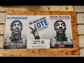 On a boarded up section of a brick wall, three posters. The outer two show Snoop Dogg's face, and the middle one shows a hand making a "V" gesture. Text of the 3 posters: 1. "I'm voting because I want to end police brutality." 2. "We need your vote. Black lives matter. Black votes matter. Register here. [QR code.]" 3. "I'm voting this election for the first time because I can't stand to see that punk in office another year." 