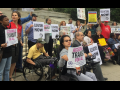 Demonstrators, some in wheelchairs and some not, stand in front of a public building. Many are holding signs: "ACCESSIBLE TRANSIT FOR ALL," "ELEVATORS NOW," "STRANDED BY CUOMO" 