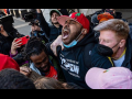 Tight crowd of people outdoors embracing and yelling in celebration. Most visible person in center, face upturned, wears a red baseball cap and a T-shirt that says The Congress of Essential Workers. Behind, many people hold up cameras capturing the moment.