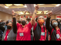 Four women in red scrubs, of various races, stand, all masked, raising their fists in the air, some clasping one another's hands. They are at the front of a large crowd in the ballroom.