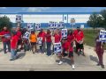 A group of Black and white men and women, most in red T-shirts, stand in front of the plant gates. Many carry blue and white "UAW on strike" picket signs.  