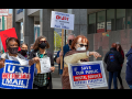 Picketers (Black and white, men and women) marching in front of a post office. Signs say "U.S. Mail Is Not for Sale," "Postmaster DeJoy Is Destroying Our Post Office," "Save Our Public Postal Service: Forever Essential." Also visible are copies of the flyer "Dump DeJoy and His 10-Year Plan."