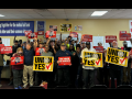A group of 33 Black and white women and men stands in a union hall, smiling and looking determined, many holding fists in the air. Many hold printed union signs. Yellow ones say "Union Yes." Red ones say "We are Alabama, we are UAW" and "No voice, no choice." Various signs on the wall behind them, including a big blue banner that says "Workers joining together for the mutual aid and protection of each other and their common interests."