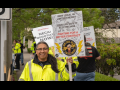 Picketers picket on a sidewalk. Person in foreground, wearing a safety-yellow jacket, smiles at the camera. This person has brown skin, glasses, hair in a ponytail, and holds a Local 46 picket sign printed with a limited energy of a fist holding bolts of lightning and the words "Your Life Safety Electricians, Fighting for a Better Contract." The sign appears to be autographed by many other strikers. Another printed sign visible in background says "On strike! NECA! Negotiate now!"