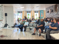 In a bright sunny room, a man in a black polo shirt and jeans stands on the left, speaking into mic, while about 30 people seated in chairs on the right listen. Most or all appear Latino/a. 