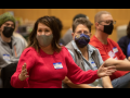 People in masks and nametags seated inside. One woman in red sweater is gesturing while speaking; others are listening.