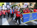 Workers march in downtown streets behind a big blue banner that says "UNITE HERE Local 2." They are wearing red T-shirts that say "Come back stronger" and some carry picket signs saying "Full service now!" or "Hotel rooms should be cleaned every day."  Most wear masks, one holds a bullhorn, and there is a child near the front of the crowd.