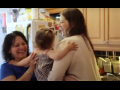 A white woman, right, hands a baby to a Latina woman, left. They are in an apartment kitchen. Both are smiling big. The baby's face is not visible.