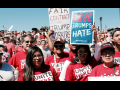 Workers in red UNITE HERE shirts pose at a huge rally. One handlettered sign says "Fair contract for Trump workers" and another printed sign says "Love Trumps Hate."