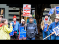 Letter carriers stand outdoors in the rain, rallying, listening to a speaker with a mic. Printed picket signs say "We deliver--how about a fair contract?" "End closed bargaining" "Harder work deserves a fair contract" and "Fair contract now."