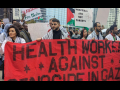 Workers stand outdoors, many wearing keffiyehs, holding a large red hand-painted banner that says "Health care workers against genocide in Gaza." Signs visible in crowd behind them say "Hospitals are not targets," "Free Palestine," and something ending in "slaughter." 
