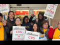 Eight people, varied in race and gender, pose together in front of a CVS store. Some wear UFCW yellow shirts and most carry picket signs that say "UFCW Local 770: CVS employees on unfair labor practices strike, please respect the picket line" in English and Spanish.