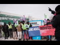 Workers, most of them Black, bundled against the cold, stand in front of an Amazon delivery facility sign. Their banner says, in French, "Without us, nothing is free" and the name of the union. Many hold CSN flags, and smile resolutely. On the right, a figure viewed from back silhouette speaks to them, one hand pointing the way forward.