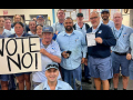 Seventeen uniformed letter carriers pose together in a post office, holding a big handmade "VOTE NO!" sign.