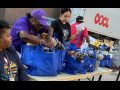 Three women work around a table with blue cloth grocery bags, outdoors. A man in a red T-shirt walks behind them. A truck is also visible.