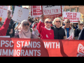 Workers, mostly women, carry a big red banner with white letters: "PSC stands with immigrants." They are in a big crowd. Some also carry picket signs, some printed and some handwritten, with messages (some partially visible) including "Dictators don't belong here," "America? Fight for it!" "Oranges don't belong in the White House," "Stop the war on immigrants," and "Stop the coup." One woman wears a hijab.