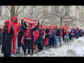 A long procession of strikers bundled up against the cold, most wearing bright red NYSNA hats or scarves, packs a sidewalk lined with piles of shoveled snow. The group is diverse in race and gender. Many are smiling. A woman in the foreground holds up a red "In Solidarity" banner.
