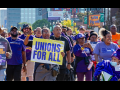 Workers march through a Detroit street, part of a Labor Day march. The people visible are mostly wearing purple SEIU T-shirts, many are Black and one holds a big printed sign readgin "Unions for all" on a yellow background. Another printed sign says "Families first, not billionaires. Not one more cut." One woman in a "Michigan home care workers" shirt pushes another in a wheelchair, also in purple gear.