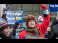 A crowd of federal workers stand outside, bundled against cold, carrying printed AFGE signs that say "EPA employees deserve respect" and "Stop the shutdown." In the foreground are a Black woman and a white woman, the latter raising a red-gloved fist in the air, both chanting or singing.