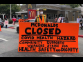 masked worker in front of mcdonald's holds big orange sign: "MCDONALD'S CLOSED, COVID HEALTH HAZARD, COMMUNITY SUPPORTS WORKERS STRIKE FOR HEALTH, SAFETY AND DIGNITY"