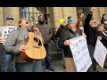 A group of people stands outside a building, singing. Edwin, a young man with guitar, has his head leaned back shouting into the sky. Several other people have fists in the air, one wears a big smile, and some hold big paper on which lyrics are written out: "Sign me up for the union, bring me my union card, We can work better with decent pay, health care, childcare, and more of a say..."