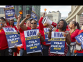 Workers in red shirts chant. Most are Black women. Many fists in air, one blowing whistle, one speaking into bullhorn. Printed signs say "UAW Stand Up, COLA and Fair Pay Now" 