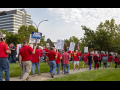 Workers in red shirts picket outside. Many of the picket signs are white with UAW logo, Local 412, and the words "Stellantis Bargaining in Bad Faith."