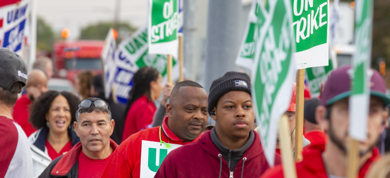 A group of UAW members march holding picket signs that say "UAW On Strike."
