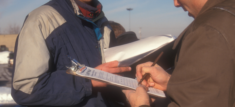 A closeup of a person’s hand signing paper on a clipboard. The person is wearing a brown uniform, possibly from UPS. Someone else is standing by in a blue and silver jacket.