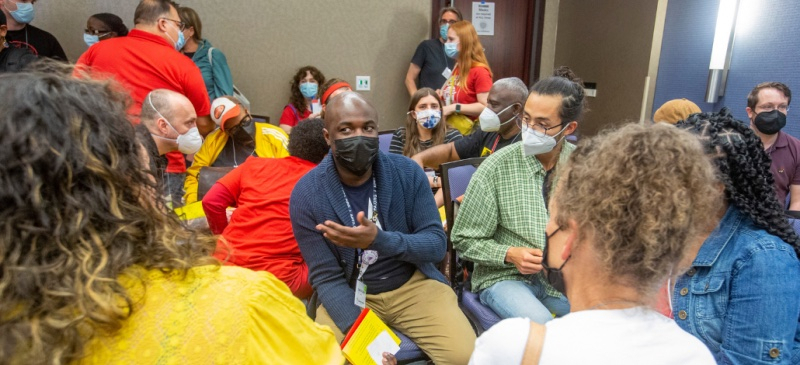 A diverse crowd of masked people talks in small groups in a conference room.