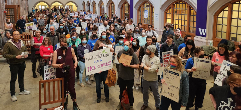 A group of 80 people with signs stand inside a collegiate-looking building. A woman in front holds a sign saying “Bioengineering for a Fair Contract.”