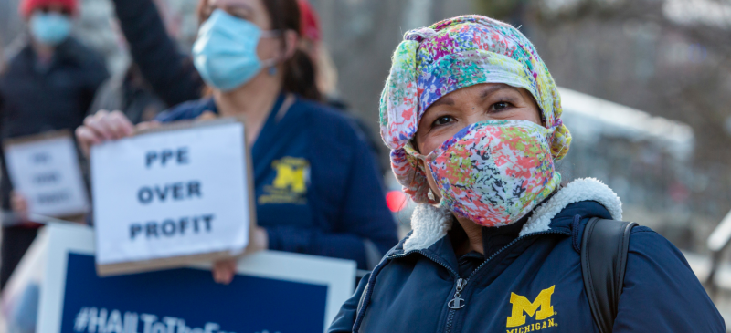 Health care workers outdoors in U of Michigan Medical Center coats. Person in front wears a colorful mask and smiles right into the camera. Behind, person holds signs "PPE OVER PROFIT" and "#HAILToTheFrontLine"