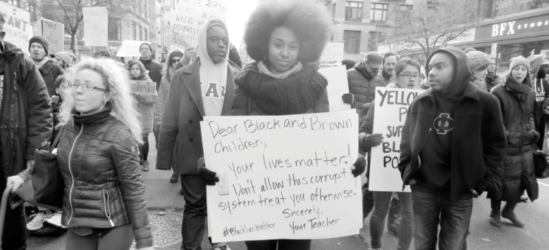 Black-and-white photo shows people marching in a New York City street, carrying signs. In the center, looking directly into the camera with a slight smile, is a Black woman with a natural hairstyle, carrying a hand-lettered sign: "Dear Black and Brown children, Your lives matter! Don't allow this corrupt system treat you otherwise! Sincerely, Your Teacher. #BlackLivesMatter"