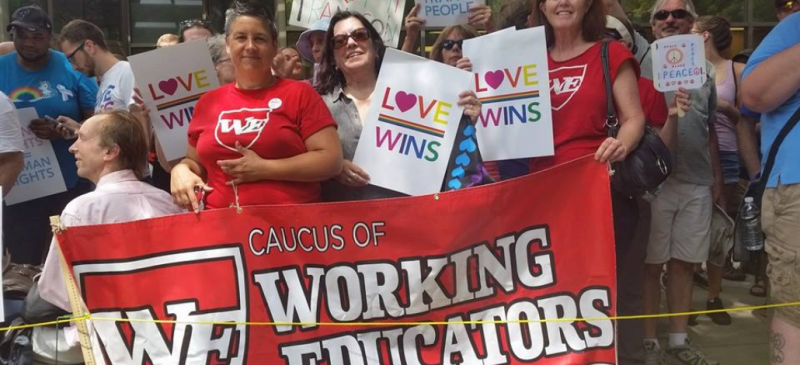 Three women mostly in red shirts looking at the camera hold signs saying “Love Wins,” and a red banner with outlined white lettering saying “Caucus of Working Educators.” 