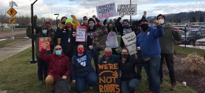 A group of Amazon solidarity activists pose for a photo with their fist raised. Some hold signs that read "Put Workers First," "Amazon Workers Need a Union," and "Solidarity Forever."