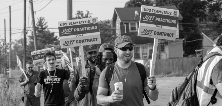 Black and white photo shows Black and white men picketing in a residential area with "UPS Teamsters: Just Practicing for a Just Contract" signs