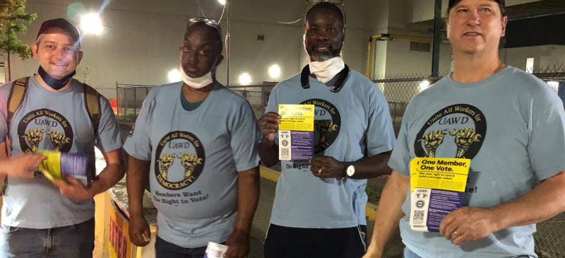 Four men of various races in blue UAWD shirts face the camera holding one-member one-vote fliers.