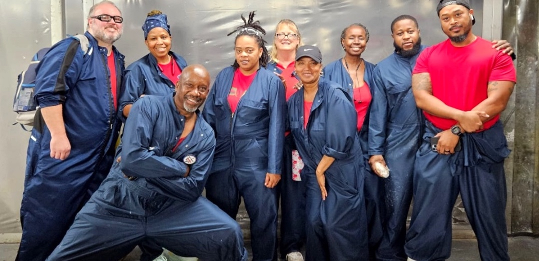 Nine workers (most of them Black, a mix of women and men) pose in red T-shirts and blue work overalls, apparently inside the plant. Their pose evinces camaraderie and confidence. One man in the front has a particularly dynamic pose with a lunge, crossed arms, and a big smile.