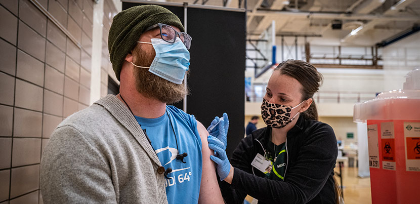 Woman on right in mask administering covid vaccine to man in mask and hat on left.