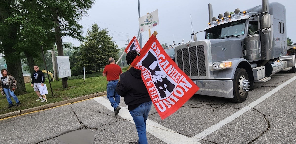 workers cross the road carrying UE flags in front of a grey semi tractor-trailer