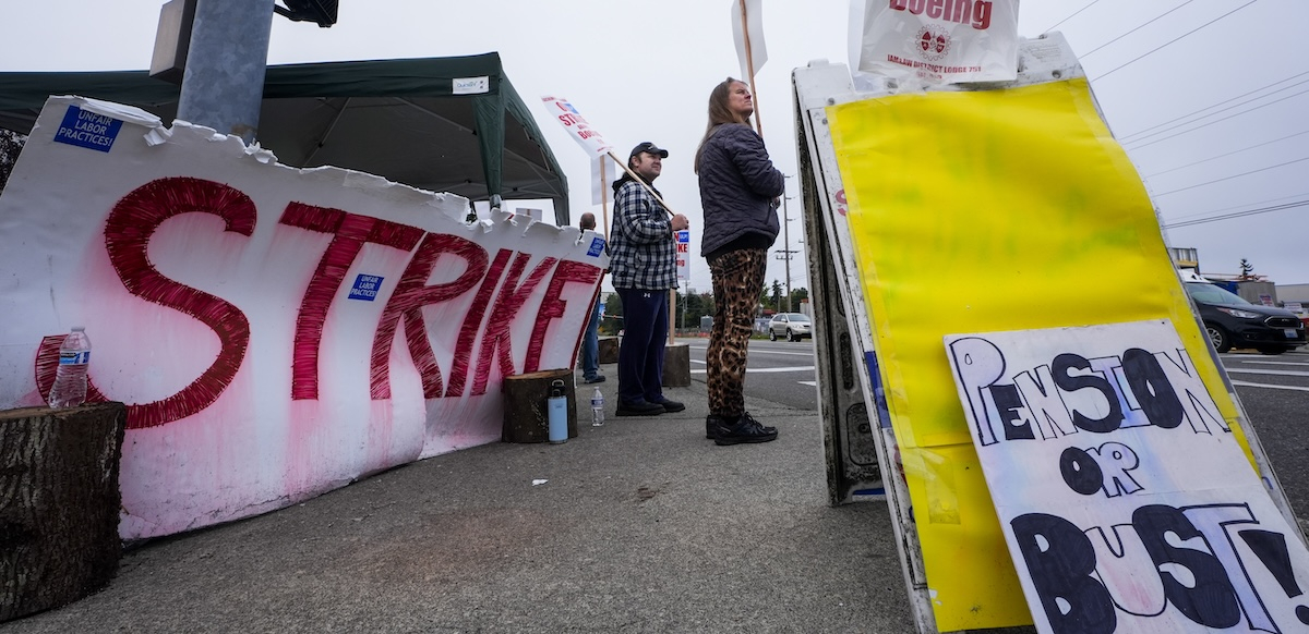 Two workers staff a picket line with a giant red ‘strike’ sign and a “Pension or Bust” sign.