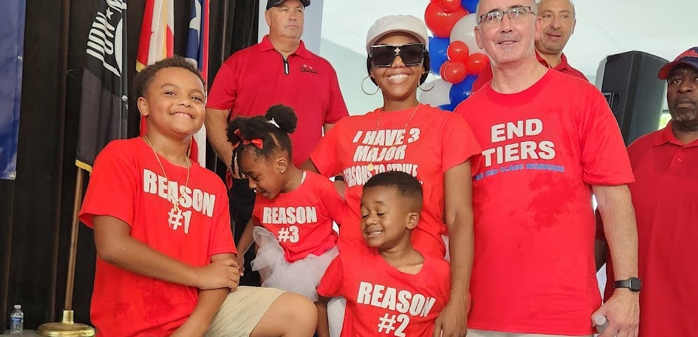 A young Black woman and her three children, all in red shirts, pose with UAW president Shawn Fain, a white man, also in a red shirt.