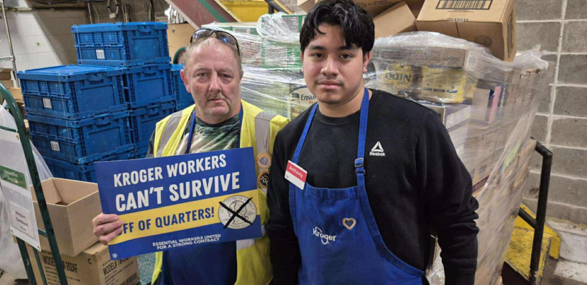 Two Kroger workers stand in front of pallets. One holds a sign that says "Kroger workers can't survive off of quarters."