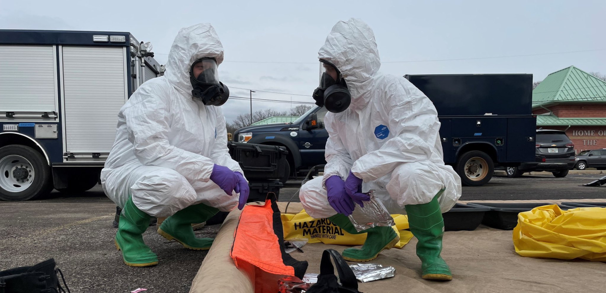 Two people in respirator masks and white hazardous materials suits squat and look at each other over a bright orange bag labeled "hazardous"
