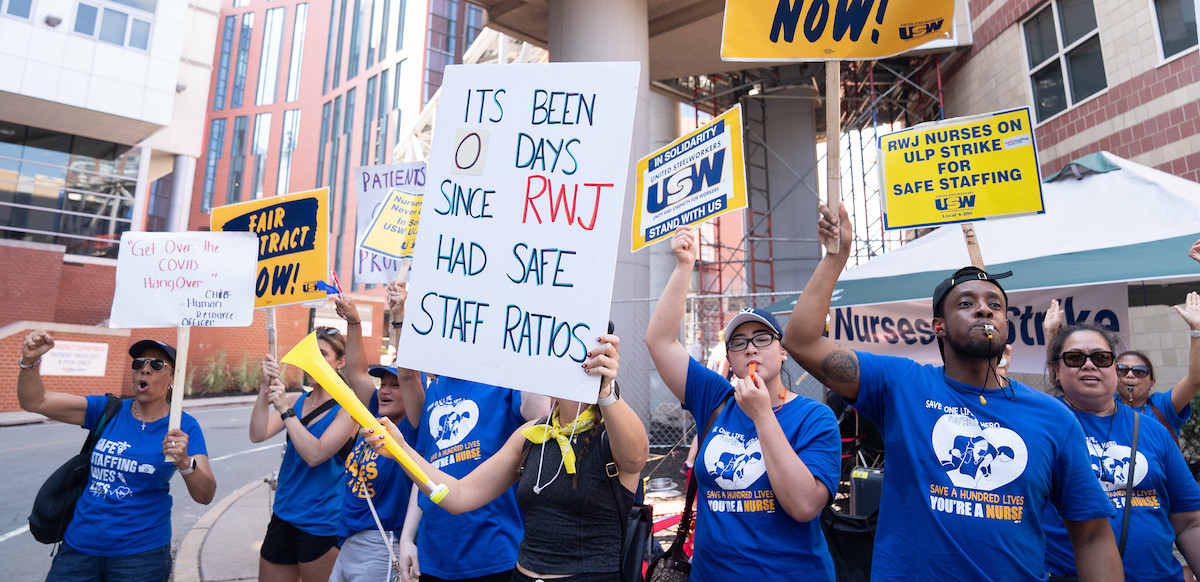 Nurses in blue T-shirts blow whistles and air horns outside a red-brick building with huge white columns.